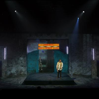 Irfan Shamji and Scott Karim standing on stage, with neon lights, racks of clothes on hangers, blue doors and a painted sign visible. Akhila Krishnan.