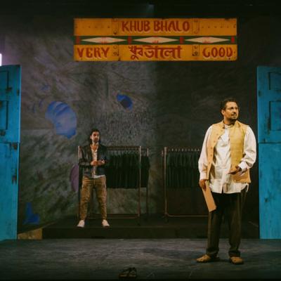 Irfan Shamji and Scott Karim standing in front of a painted sign reading 'Khub Bhalo', blue doors and racks of clothing on hangers. Isha Shah. 
