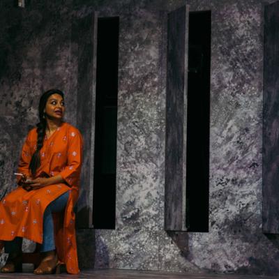 Ayesha Dharker sitting on a chair in front of a grey painted wall with vents cut out. Isha Shah.