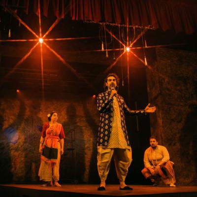 Scott Karim, Ayesha Dharker and Irfan Shamji standing on stage, with lights and a red curtain visible above. Scott Karim holds a microphone to mouth as if speaking. Isha Shah.