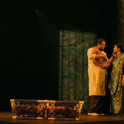 Irfan Shamji and Zainab Hasan standing on stage together, with Irfan Shamji holding a baby. A woven bassinet for a baby is on the floor next to them. Isha Shah.