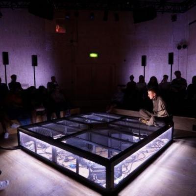 The whole audience is in low lighting, the stage is illuminated in a wash of purple lighting. In the middle, Ivan sat with his legs crossed on the stage. Photo by Anthony Lee at the Young Vic.