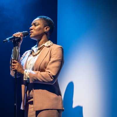 A Black woman of average height with short hair stands singing in front of a microphone. She wears a peach coloured suit.