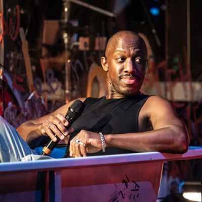 A Black man of average height with short afro hair sits in a bathtub, holding a microphone. He wears a Black vest and denim jeans.