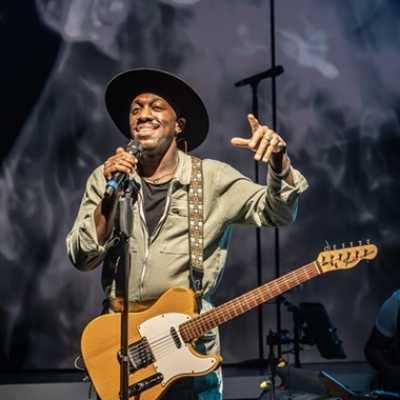 A Black man of average height stands singing in front of a microphone. He holds one hand up, gesturing. He wears a wide-brimmed hat and a boilersuit, and has a guitar on him. 