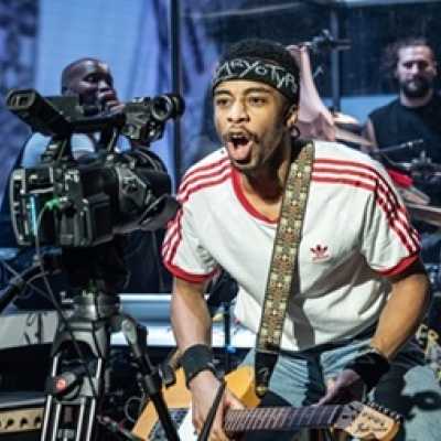 An average height, slim, Afro-Caribbean man wearing a red and white top with a band tied around his head. He shouts into a camera just in front of him, and wears an electric guitar. In the background there is a band.