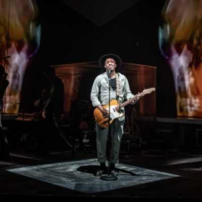 A Black man of average height stands singing in front of a microphone. He wears a wide-brimmed hat and a boilersuit, and has a guitar on him. Images are visible on the wall behind him.