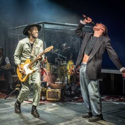 A Black man of average height, wearing a wide-brimmed hat and a boilersuit, plays electric guitar. Near him, a man with light-brown skin, also of average height, sings into a microphone. He wears a cropped suit jacket, a cropped top and jeans. Behind them is a box with glass walls with graffiti on it. 