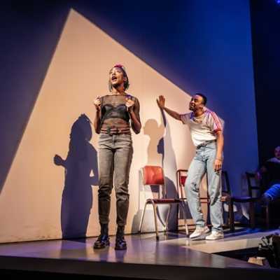 A tall Black woman with short black cropped hair stands, wearing a mesh t-shirt and jeans, with a scrunchie in her hair. Behind her, an average height, slim,  Afro-Caribbean man wearing a red and white top leans against a wall.