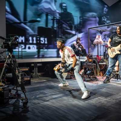 An average height, slim, Afro-Caribbean man wearing a red and white top and jeans, and converse, with a band tied around his head. He holds an electric guitar and talks towards a camera off to the left side of the image. Next to him a mixed race girl with long braids stands playing an electric guitar, she wears a black t-shirt, jeans and doc martens.