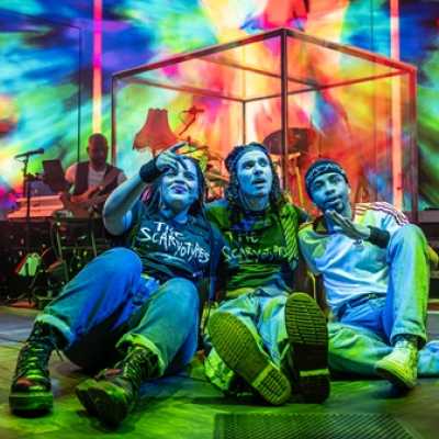 Three people sit on the floor in front of a large video screen showing a psychelic, tie-dye type pattern. The pattern is multi-coloured. In between them and the screen a drum kit is visible. 