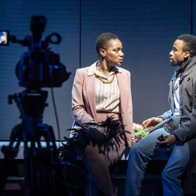 Two people sit next to each other, facing each other. One is a Black woman of average height with short hair, wearing a peach coloured suit. The other is an average height, slim, Afro-Caribbean man wearing a dark trench coat with a turned-up collar. In the foreground, a camera is visible.