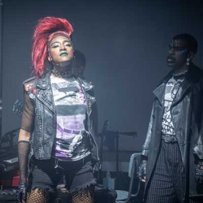 A tall Black woman with red hair styled into a loose mohawk wearing a leather sleeveless coat, a black mesh t-shirt, short and fishnet tights. Next to her is a Black man with make-up on his face, wearing a leather coat.