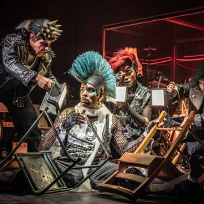 A Black man with a blue mohawk, wearing a white vest, pin-striped trousers, braces and a fishnet under-shirt sits on the floor with a glum expression on his face. Two upturned chairs are on the floor next to him. Three people kneel around him, holding up lights.