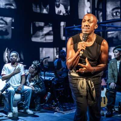 A Black man of average height sings into a microphone. Behind him an average height, slim, Afro-Caribbean man sits on a toilet with his hand clasped to his chest. Three people with brightly coloured mohawks, mesh tops and tights and leather jackets kneel around him, looking up at him. Behind them are two large walls of retro tv screens, each depicting a different image, some are of his face. On the other side of the stage a mixed race girl with long braids wearing a beret and green jacket looks over at the 