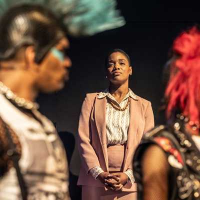 A Black woman of average height with short hair, wearing a peach coloured suit, stands between a Black man and tall Black woman in the foreground, with their hair styled into mohawks, wearing black mesh shirts and sleeveless jackets.