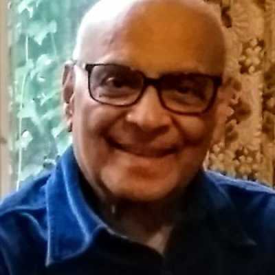 An elderly man, with a bald head and black glasses, smiles at the camera