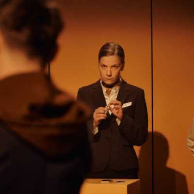 A white woman with tied-back blonde hair, wearing a dark suit, stands next to a small, dark-haired woman with mid-length hair tied back into a ponytail. The woman in the dark suit holds something up to her finger. In the foreground of the image, slightly out of focus, is a Caucasian man with sandy hair.