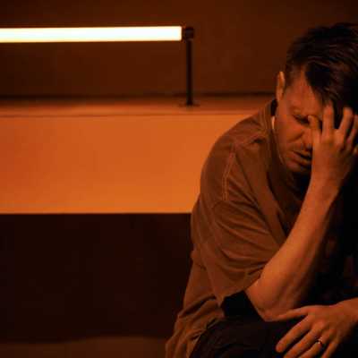 In an orange room bathed in orange light, a Caucasian man with sandy hair sits with his hand up to his face.