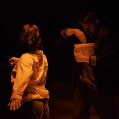 A Caucasian man with sandy hair dips his hand into a bucket of custard; a small, dark-haired woman facing away from the camera is about to throw custard at him.