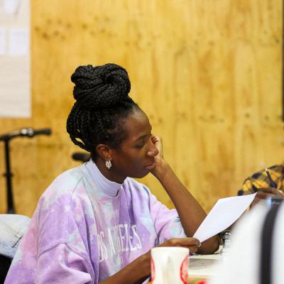 Actress Danielle Kassaraté sits at a table holding a script and is looking down at it while Kassaraté's left hand rests on Kassaraté's chin. 
