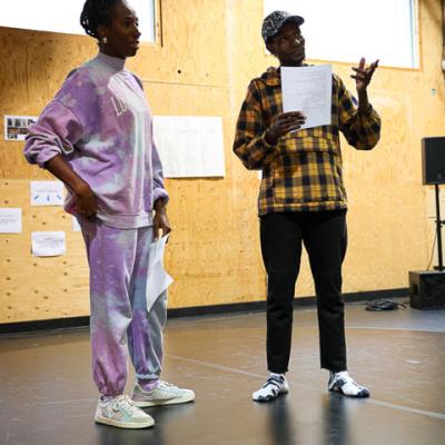 Actor John Rwothomach and Actress Danielle Kassaraté stand on stage with scripts in hand looking towards the audience. 