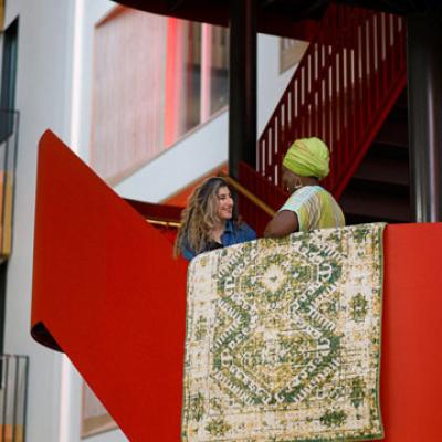 Two people standing on a red staircase, with a rug hung over one of the bannisters