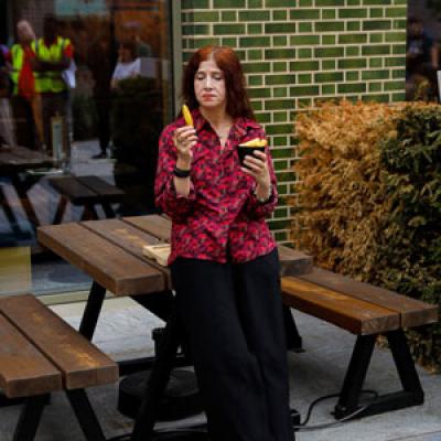 A person outside, leaning against a picnic table, eating chips