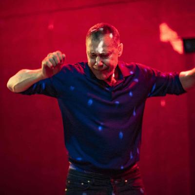 Hans Kesting is surrounded by red light and wearing a blue jumper, holding hands up as if dancing; fragments of light reflecting off of a disco ball are visible.