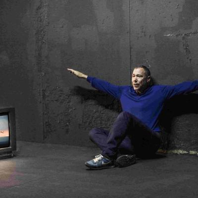 Hans Kesting is sitting down on the floor with arms outstretched, mimicking a scene visible on a nearby television from Titanic (1997) where Jack and Rose stand at the helm of a ship with both their arms spread out.