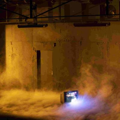 Stage bathed in orange light, with the floor covered in a thick haze. A television on the floor displays an image of a ship, and Hans Kesting leans against the wall.