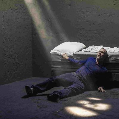 Hans Kesting lies on the floor, leaning against a bed. Kesting is covered in dirt, and rays of light coming from a source out of frame illuminate Kesting.