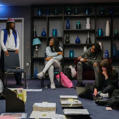 A group of people sitting in a room with grey walls, blue carpet and vases on shelves in the background. Some people are sitting on chairs, talking to each other, and others sit on the floor, looking at documents or using laptops.