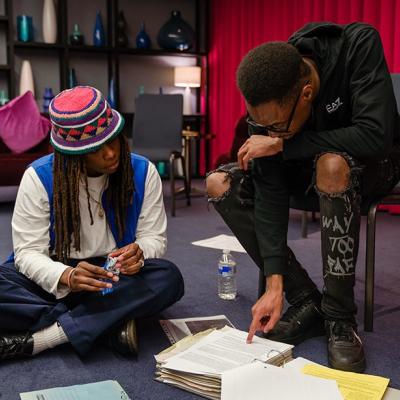 Two people looking at a folder of documents on the floor, with one person crouching on the floor and one person sitting on a chair