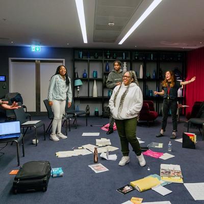 A group of people standing in a room with grey walls, blue carpet and vases on shelves in the background. On the floor there are pieces of paper and water bottles.