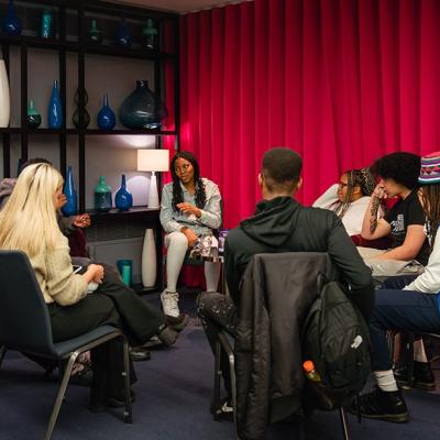 A group of people sitting in chairs in a circle, in a room with grey walls with blue vases displayed on shelves, and a red curtain pulled over one of the walls.