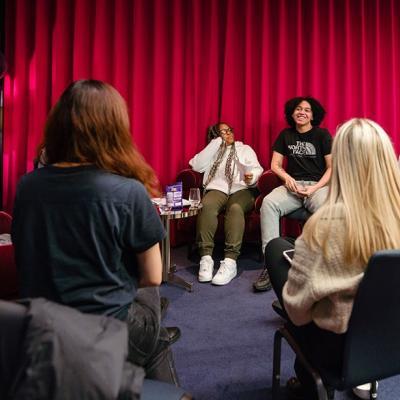 A group of people sitting in chairs in a circle, smiling and laughing as they talk to each other, in front of a wall with a red curtain pulled across it