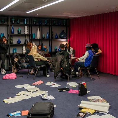 A group of people sitting in chairs in a circle, in the corner of a large room with with grey walls, blue carpet and vases on shelves in the background, and a red curtain. There are pieces of paper, files and bags on the floor.