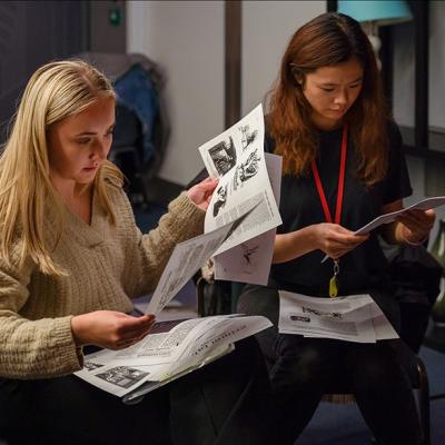 Two people sit on chairs as they look through a pile of papers with text and black and white images and diagrams on.