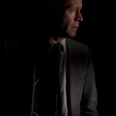 Portrait of Jonathan Cullen (Man) standing in almost complete darkness with his face, suit and tie dimly lit looking intently across the stage in Winter at the Young Vic © Ellie Kurttz