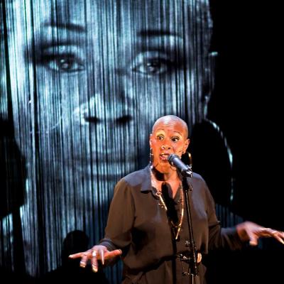 Josette Bushell-Mingo in Nina with beautiful fringed curtains and a larger projection of her face behind her. Photo by Simon Annand