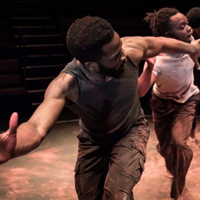 The three cast members stand in a line with their arms stretched out. The lighting is dark with an orange hue. Sope leads the group followed by Jonathan then Anthony. Photo by Marc Brenner.