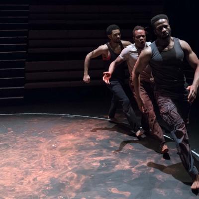 The three cast members are marching angrily around the inside of a chalk circle drawn on the floor. Dry red paint is smeared around the floor. Photo by Marc Brenner