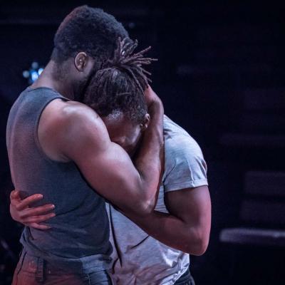 Sope and Jonathan are embraced in a hug. Neither of their faces are visible but it looks mornful. The lighting had a blue hue. Photo by Marc Brenner.