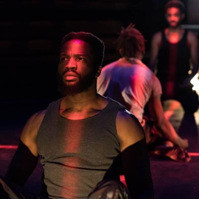 Sope (Ogun Size) sits on the floor. A line of red light is illuminating his face. Behind and out of focus you can see the characters Elegba and Oshoosi. Photo by Marc Brenner.