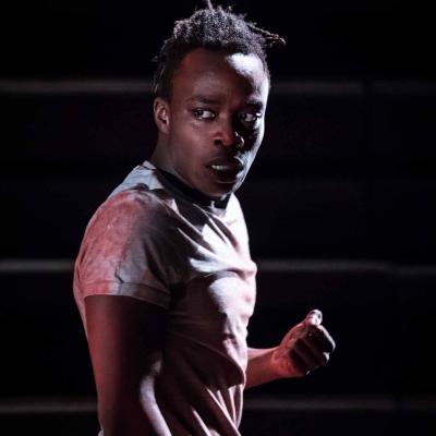 Jonathan Ajayi (Oshoosi Size) is standing in portrait looking worried. He is wearing a grey t-shirt that is smeared with dry red paint. Photo by Marc Brenner.