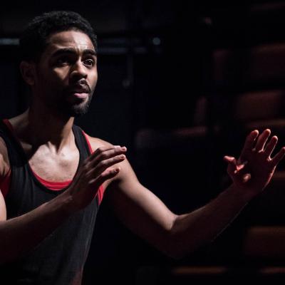 Anthony Welsh (Elegba) is wearing a black vest with a red vest below. He looks like he is pleading to somebody with his arms outstretched. Photo by Marc Brenner.