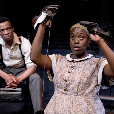 Production photo of Things of Dry Hours. The set is dimly lit with dark wooden floorboards and a dirty wooden slated backdrop. Washing hangs in the background. The actors wear basic 1900s era working dress.