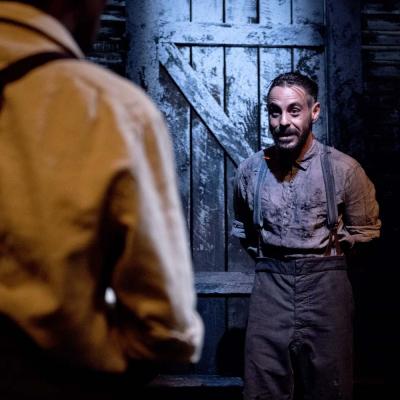 Production photo of Things of Dry Hours. The set is dimly lit with dark wooden floorboards and a dirty wooden slated backdrop. Washing hangs in the background. The actors wear basic 1900s era working dress.