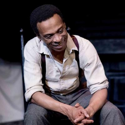 Production photo of Things of Dry Hours. The set is dimly lit with dark wooden floorboards and a dirty wooden slated backdrop. Washing hangs in the background. The actors wear basic 1900s era working dress.